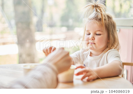 Cute little girl is eating ice cream. A beautiful charming baby girl sitting at a table in a cafe near the window. Children's cafe. The interior of an ice cream cafe. 104086019