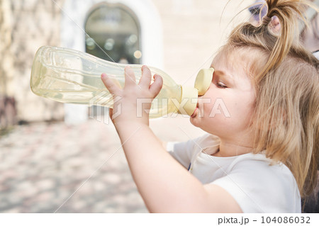 a charming little girl drinks water from a sports bottle in a city park. a close-up portrait of a child who drinks from a bottle on a warm summer day. 104086032
