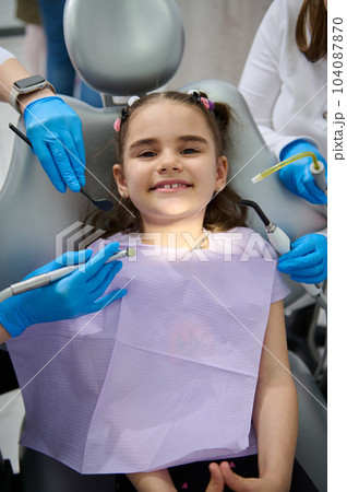 Top view. Hands of a dentist and assistant hold sterile dental instruments - dental mirror, probe, drill and saliva ejector above a smiling little patient at appointment in pediatric dentistry clinic 104087870
