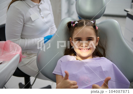 Smiling little girl at dentist appointment, sitting on dental chair, showing thumbs up after preventive dental check up in modern children's dental clinic. Pediatric dentistry. Prevention of caries Smiling little girl at dentist appointment, sitting on dental chair, showing thumbs up after preventive dental check up in modern children's dental clinic. Pediatric dentistry. Prevention of caries 104087871