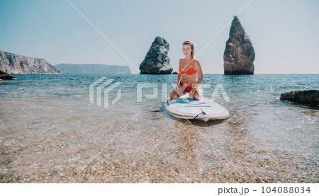 Woman sea sup. Close up portrait of happy young caucasian woman with blond hair looking at camera and smiling. Cute woman portrait in red bikini posing on sup board in the sea 104088034
