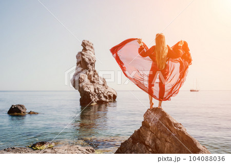 Woman travel sea. Young Happy woman in a long red dress posing on a beach near the sea on background of volcanic rocks, like in Iceland, sharing travel adventure journey 104088036