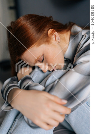 Close-up vertical portrait of sad millennial redhead female sitting on bed embrace knees lost in bad pessimistic thoughts suffer alone with closed eyes. Stressed young woman feel lonely desperate 104089589