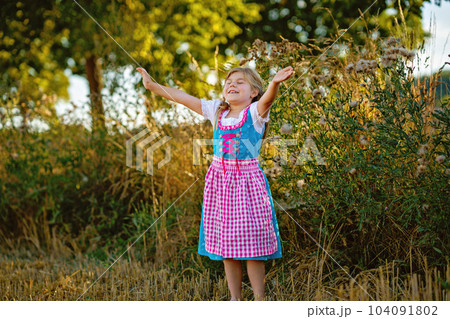 Cute little kid girl in traditional Bavarian costume in wheat field. Happy child with hay bale during Oktoberfest in Munich. Preschool girl play at hay bales during summer harvest time in Germany. Cute little kid girl in traditional Bavarian costume in wheat field. Happy child with hay bale during Oktoberfest in Munich. Preschool girl play at hay bales during summer harvest time in Germany. 104091802