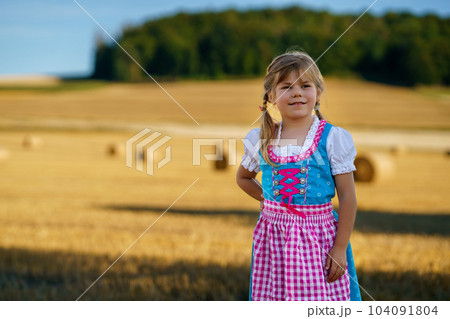 Cute little kid girl in traditional Bavarian costume in wheat field. Happy child with hay bale during Oktoberfest in Munich. Preschool girl play at hay bales during summer harvest time in Germany. 104091804