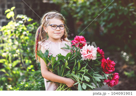 Cute adorable little preschool girl with huge bouquet of blossoming red and pink peony flowers. Portrait of smiling preschool child in domestic garden on warm spring or summer day. Summertime. Cute adorable little preschool girl with huge bouquet of blossoming red and pink peony flowers. Portrait of smiling preschool child in domestic garden on warm spring or summer day. Summertime. 104091901