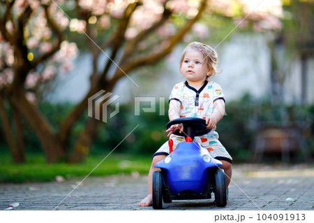 Cute little baby girl playing with blue small toy car in garden of home or nursery. Adorable beautiful toddler child with blossoming magnolia on background 104091913