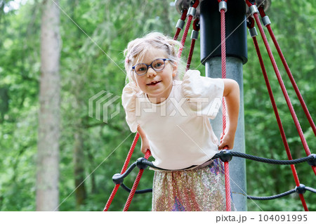 Adorable happy little girl is climbing on an outdoor playground on a summer day. Blonde preschool child climbs to the top of the game complex 104091995