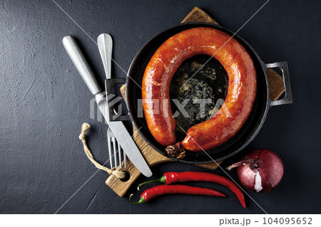 Fried sausage rolled into a ring on a cast iron pan with cutlery, a wooden cutting board, hot peppers and red onions on a black stone background 104095652
