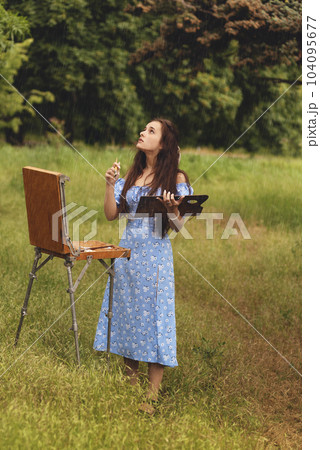 Beautiful young woman is relaxing while painting an art canvas outdoors in the park at rain. Beautiful young woman is relaxing while painting an art canvas outdoors in the park at rain. 104095677