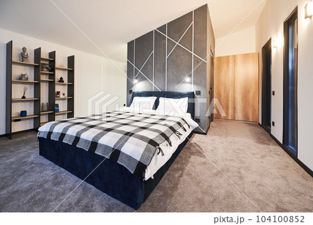 Simply furnished sleeping area with big bed and wooden shelves with buddhist decoration at home. Side view of dark bed covered with plaid bedspread against gray wall in bedroom. Concept of interior. Simply furnished sleeping area with big bed and wooden shelves with buddhist decoration at home. Side view of dark bed covered with plaid bedspread against gray wall in bedroom. Concept of interior. 104100852