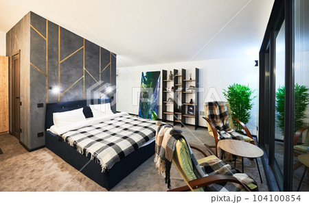 Cozy modern furnished chalet room with reflection in large panoramic window at twilight. Side view of illuminated bedroom indoors. Concept of interior. 104100854
