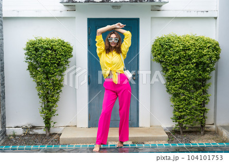 Stylish fit fashion women in bright pink wide leg pants and yellow shirt holding bag trendy mint sunglasses posing at luxury tropical villa by pool outdoor natural day light 104101753