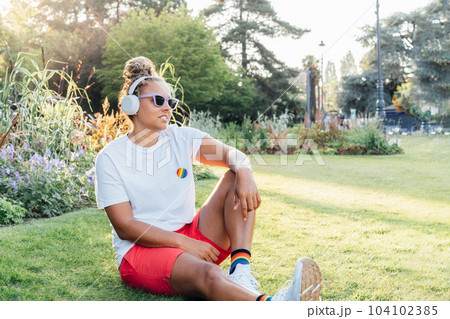 Mixed race young woman with rainbow badge, symbol of LGBTQ social community listening music with wireless headphones while relax in park. Personal Coming out, free lifestyle. Celebrating Pride month 104102385