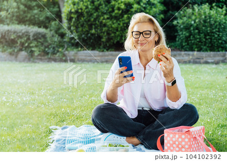 Middle aged caucasian woman sitting on green lawn, eating healthy lunch and using phone. Having picnic during break or after work. Balanced diet lunch box with salad and sanwich. Healthy eating habit. Middle aged caucasian woman sitting on green lawn, eating healthy lunch and using phone. Having picnic during break or after work. Balanced diet lunch box with salad and sanwich. Healthy eating habit. 104102392