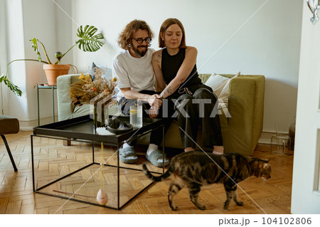 Lovely couple sitting on sofa at living room and looking on his domestic cat. Spending time together Lovely couple sitting on sofa at living room and looking on his domestic cat. Spending time together 104102806