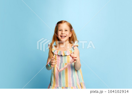 Portrait of beautiful, happy, little girl, child in multicolored striped dress posing with smile and laugh against blue studio background 104102914