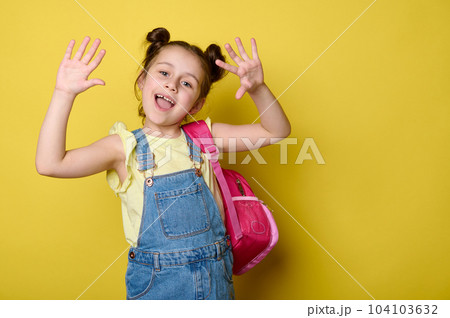 Cheerful preschooler girl showing at camera her hands palms up, smiling, enjoying the start of new school year, posing with pink backpack, dressed in blue denim dress, isolated over yellow background Cheerful preschooler girl showing at camera her hands palms up, smiling, enjoying the start of new school year, posing with pink backpack, dressed in blue denim dress, isolated over yellow background 104103632