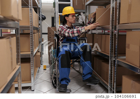 Young asian warehouse worker in wheelchair taking parcel from shelf. Storehouse order picker with physical disability searching cardboard box while working in storage room Young asian warehouse worker in wheelchair taking parcel from shelf. Storehouse order picker with physical disability searching cardboard box while working in storage room 104106691