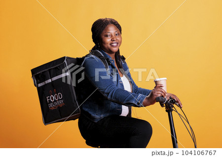 Portrait of restaurant employee courier holding cup of coffee while delivering takeaway food order to clients using bike as transportation. Pizzeria worker standing in studio with yellow background. 104106767