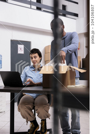 Warehouse worker putting client order in cardboard box wrapping with bubble wrap while manager checking package shipping details on laptop computer. Distribution center fulfillment company Warehouse worker putting client order in cardboard box wrapping with bubble wrap while manager checking package shipping details on laptop computer. Distribution center fulfillment company 104106807