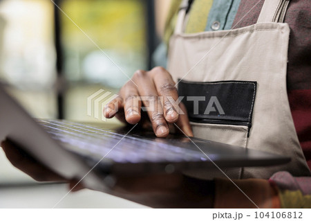 Storehouse worker checking customer order details on laptop computer, preparing goods for shipping in storage room. African american employee analyzing merchandise logistics in warehouse. Close up Storehouse worker checking customer order details on laptop computer, preparing goods for shipping in storage room. African american employee analyzing merchandise logistics in warehouse. Close up 104106812