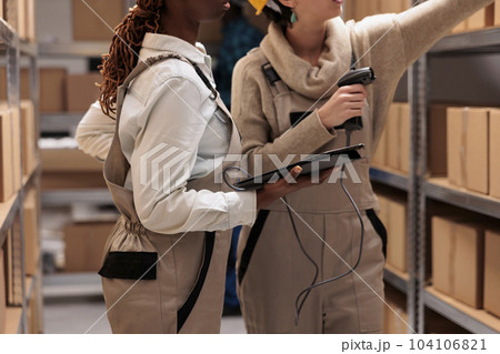 Storehouse colleagues reaching for cardboard box on shelf to scan barcode. Warehouse operators managing received parcels registration with bar code scanner and digital tablet software Storehouse colleagues reaching for cardboard box on shelf to scan barcode. Warehouse operators managing received parcels registration with bar code scanner and digital tablet software 104106821