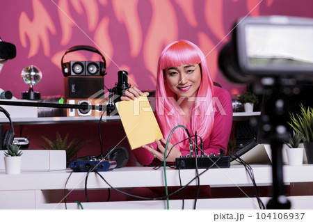 Influencer with pink hair advertising notebook in front of camera recording vlog while presenting school supplies review using professional vlogging equipment. Asian creator filming podcast Influencer with pink hair advertising notebook in front of camera recording vlog while presenting school supplies review using professional vlogging equipment. Asian creator filming podcast 104106937