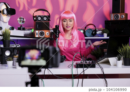 Happy vlogger with pink hair recording podcast while talking with remote subscribers in vlogging studio, posting content on social media. Influencer broadcasting vlog using filming equipment 104106990