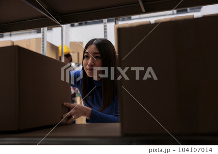 Distribution center worker taking cardboard box from rack to prepare goods for dispatching. Asian woman order picker searching carton package on shelf in industrial warehouse Distribution center worker taking cardboard box from rack to prepare goods for dispatching. Asian woman order picker searching carton package on shelf in industrial warehouse 104107014