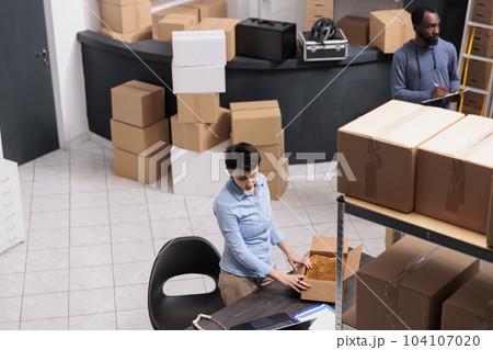 Top view of employee standing at desk in warehouse while preparing client order putting clothes in carton box after checking delivery detalies. Diverse team working in distribution center 104107020