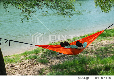 A woman in a cap lies in a hammock on the river bank 104107410