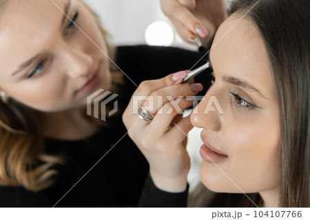 The photo captures an experienced makeup artist perfecting the client's eyebrows. Every detail counts and the master focuses on creating perfect symmetry and close-up lines in a beauty salon. The photo captures an experienced makeup artist perfecting the client's eyebrows. Every detail counts and the master focuses on creating perfect symmetry and close-up lines in a beauty salon. 104107766