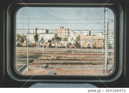 Train window view with old railway, clear blue sky, white and red brick buildings, yellow and red freight wagons, metal fence, city wires and industrial area. Pale non-contrast color correction. 104108472