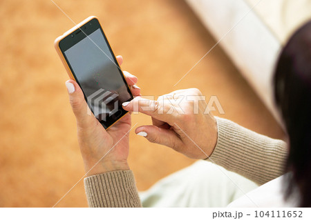 Close up of hands from middle aged woman sitting on sofa at home and using a mobile phone. 104111652
