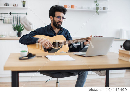 Person with guitar smiling at laptop webcam in home interior Person with guitar smiling at laptop webcam in home interior 104112490