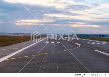 At the airport, endless runway extends into the horizon, featuring prominent aircraft markings and well-defined pathways for the safe arrival and departure of planes at sunset At the airport, endless runway extends into the horizon, featuring prominent aircraft markings and well-defined pathways for the safe arrival and departure of planes at sunset 104112583