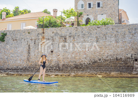 Young women Having Fun Stand Up Paddling in blue water sea in Montenegro. Against the backdrop of the Old Town of Budva. SUP 104116989