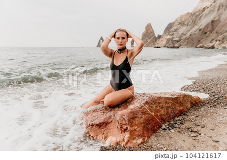 Woman travel portrait. Happy woman with long hair looking at camera and smiling. Close up portrait cute woman in black bikini posing on a red volcanic rock on the sea beach 104121617