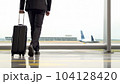 Businessman with suitcase in airport departure hall with airplane in background - AI content 104128420