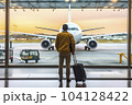 Businessman with suitcase in airport departure hall with airplane in background - AI content 104128422