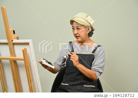 Beautiful 60s senior woman wearing apron holding palette painting picture over gray background Beautiful 60s senior woman wearing apron holding palette painting picture over gray background 104128712