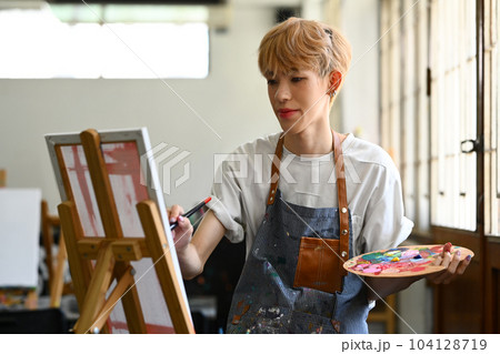 Peaceful young stylish gay man painting picture with oil paints on canvas in bright art studio. Leisure activity and art concept 104128719