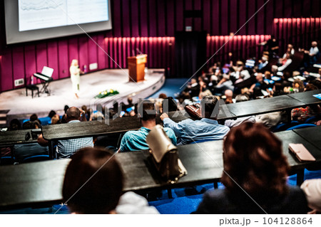 Business and entrepreneurship symposium. Female speaker giving a talk at business meeting. Audience in conference hall. Rear view of unrecognized participant in audience. 104128864