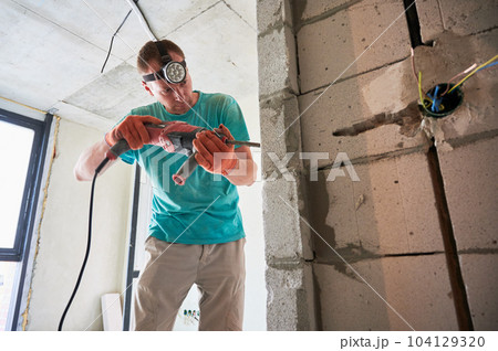 Male construction worker drilling brick wall with electric drill in apartment under renovation. Man wearing headlamp flashlight and work gloves while using professional drilling tool at home. Male construction worker drilling brick wall with electric drill in apartment under renovation. Man wearing headlamp flashlight and work gloves while using professional drilling tool at home. 104129320