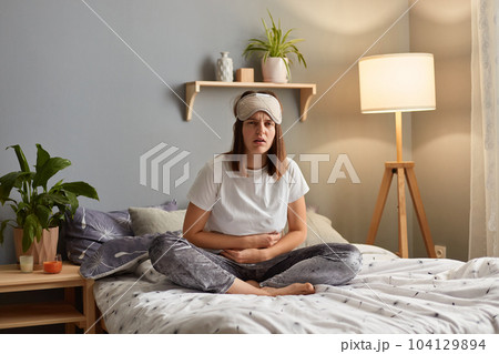 Horizontal shot of sick unhealthy sad woman wearing sleeping mask sitting on bed in cozy bedroom, having terrible strong stomachache, frowning face from pain. Horizontal shot of sick unhealthy sad woman wearing sleeping mask sitting on bed in cozy bedroom, having terrible strong stomachache, frowning face from pain. 104129894