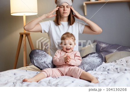 Portrait of of unhealthy woman mother wearing pajama and blindfold with her baby daughter in the bedroom sitting on bed, female suffering headache due to lack of sleep. 104130208
