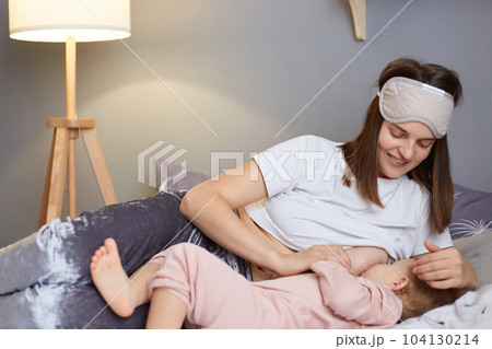 Portrait of attractive woman mother wearing blindfold breastfeeds infant baby girl while lying on home bed, smiling woman in sleeping mask feeding her toddler kid. 104130214