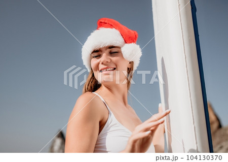 Woman sea sup. Close up portrait of happy young caucasian woman with long hair in Santa hat looking at camera and smiling. Cute woman portrait in a white bikini posing on sup board in the sea 104130370