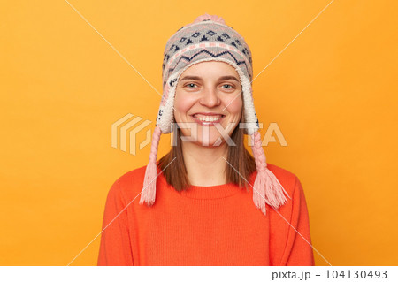 Portrait of smiling beautiful woman wearing orange jumper and knitted earflap hat standing isolated over yellow background, looking at camera with toothy smile, being in good mood. Portrait of smiling beautiful woman wearing orange jumper and knitted earflap hat standing isolated over yellow background, looking at camera with toothy smile, being in good mood. 104130493